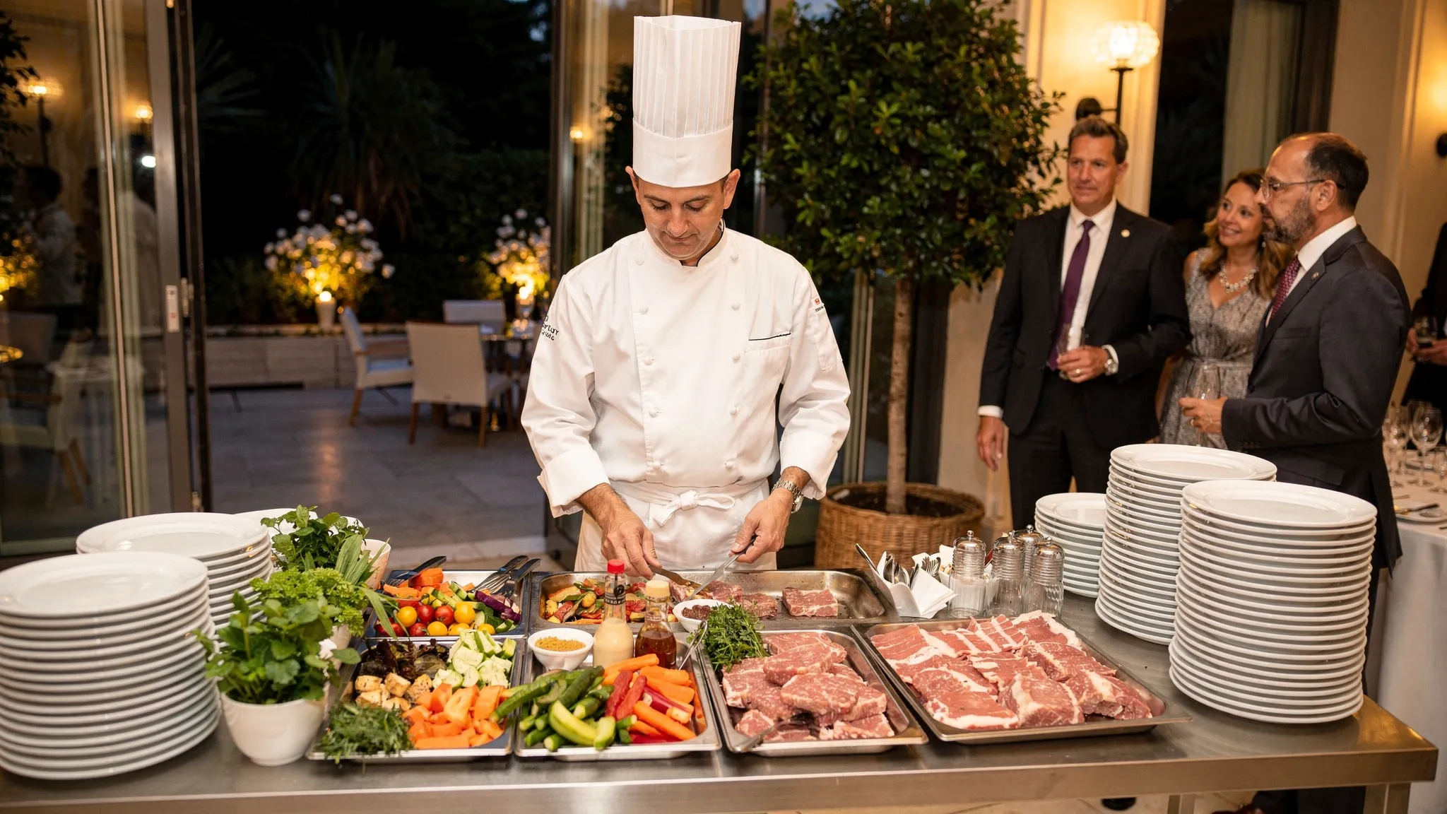 Stație de live cooking la un eveniment: chef în fața unei mese de lucru, ingrediente proaspete, farfurii pregătite pentru servire și invitați care se apropie, într-un spațiu elegant indoor-outdoor.