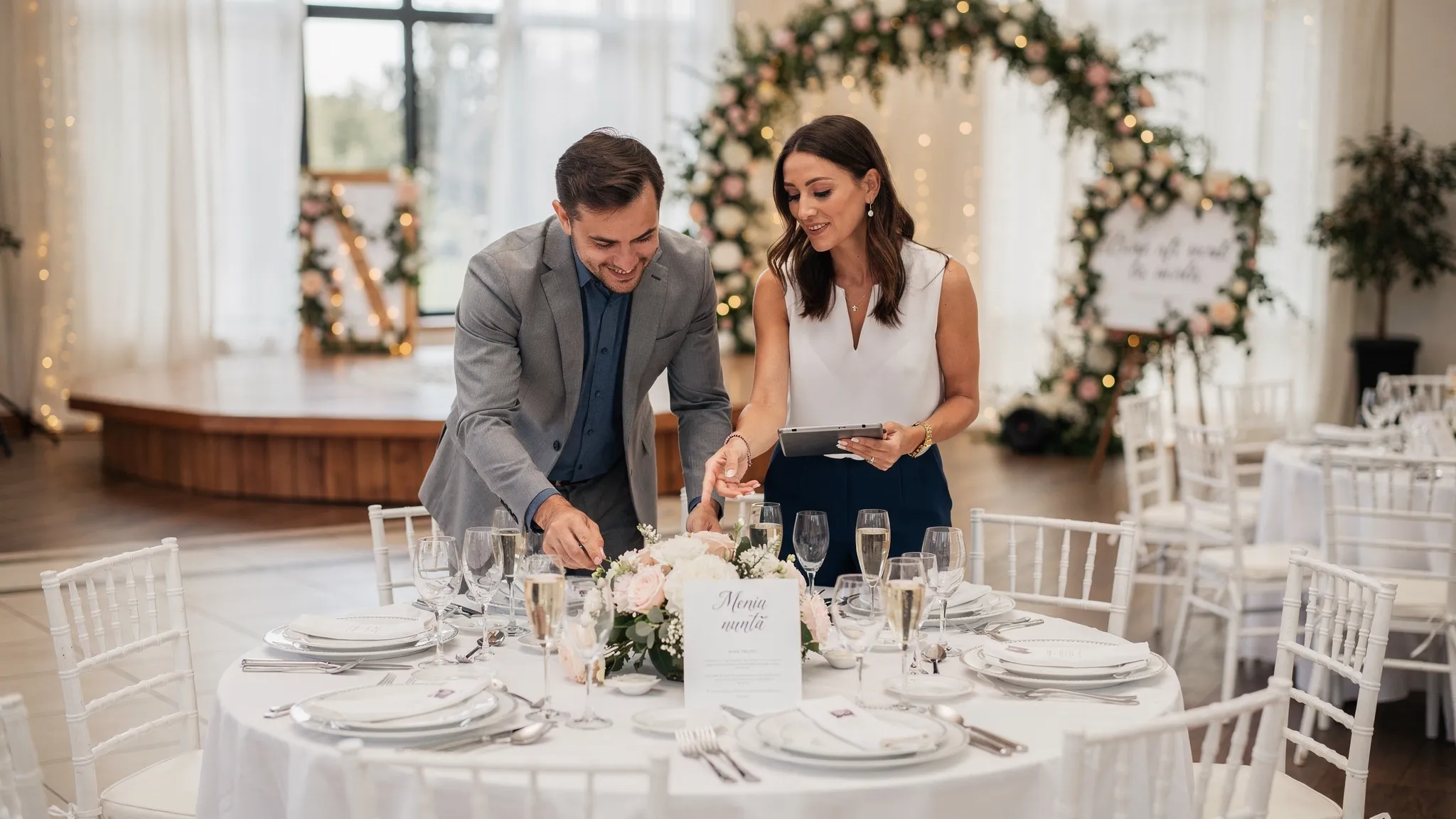 Locații nuntă București: top criterii pentru o alegere sigură 2 Un cuplu vizitează o locație de nuntă împreună cu un consultant, discutând lângă o masă aranjată festiv; în fundal se vede ringul de dans și zona de ceremonie.