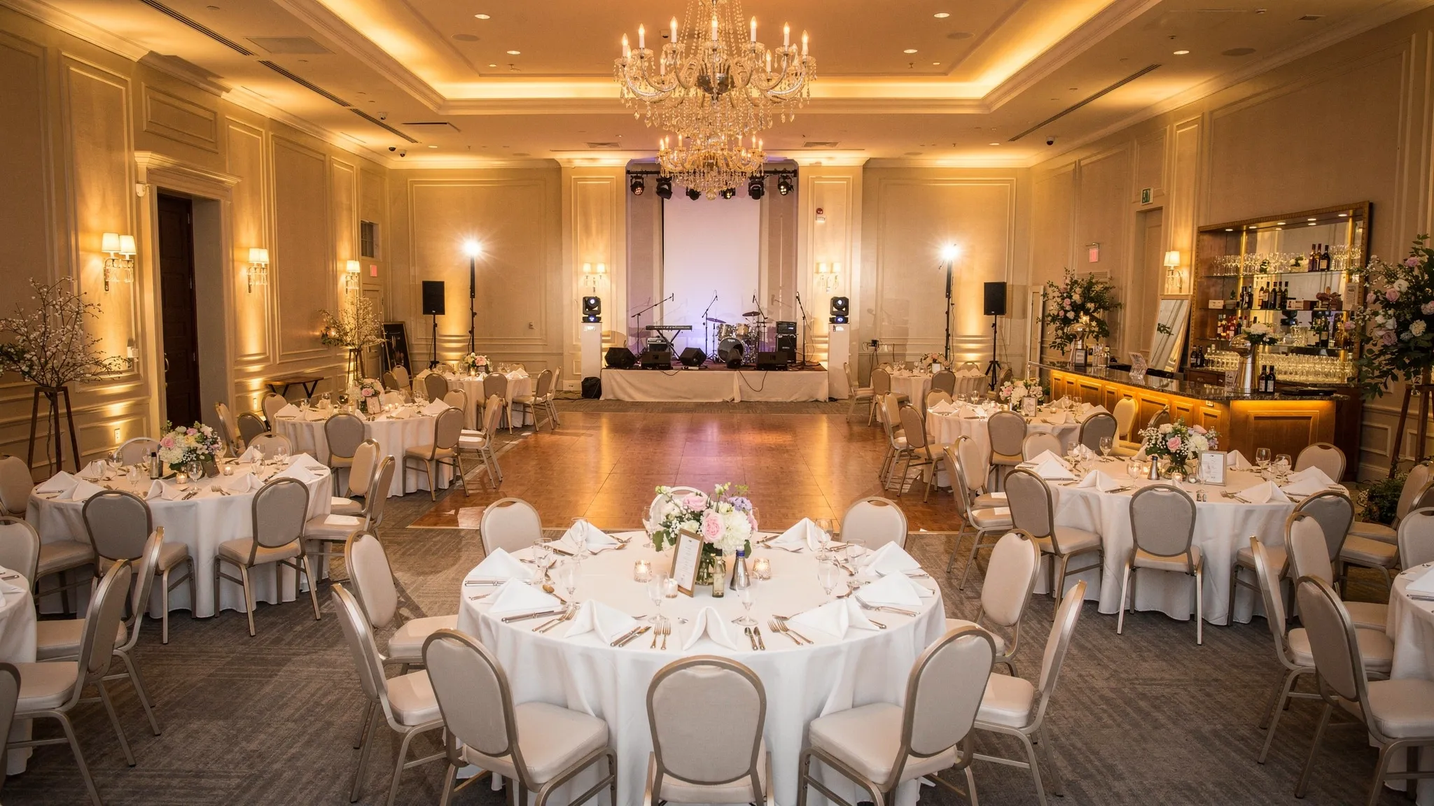 O sală elegantă de nuntă cu mese aranjate pentru invitați, ring de dans central, iluminat cald, elemente florale discrete și un spațiu clar pentru scenă și bar, sugerând o organizare premium și un flux aerisit al serii.
