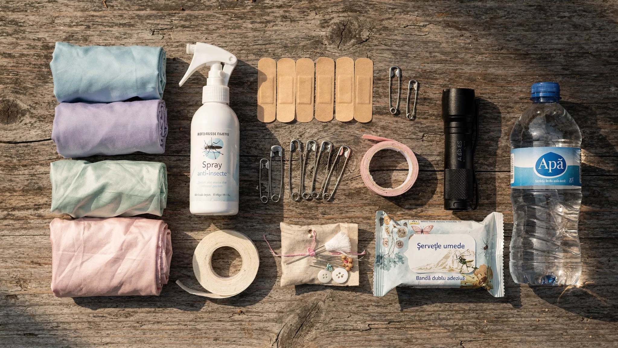 Nuntă în natură: checklist pentru o zi fără surprize 3 Flat lay cu un kit de urgență pentru nuntă în natură: pelerine de ploaie pliate, spray anti-insecte, plasturi, ace de siguranță, bandă dublu adezivă, lanternă, șervețele umede, mini trusă de cusut și o sticlă de apă, așezate pe o masă de lemn.