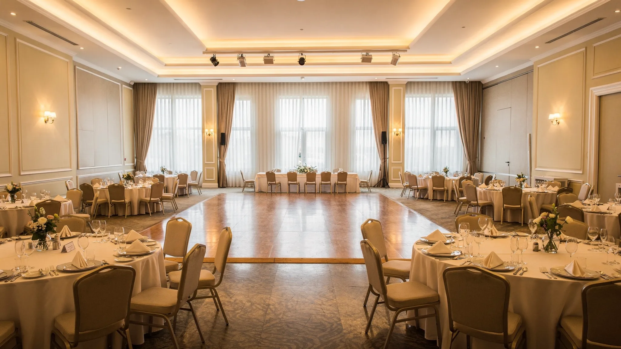 Interior de ballroom elegant pentru evenimente în București, cu tavan înalt, iluminat cald în straturi (ambiental și spoturi), mese rotunde aranjate aerisit, ring de dans central și decor floral discret.
