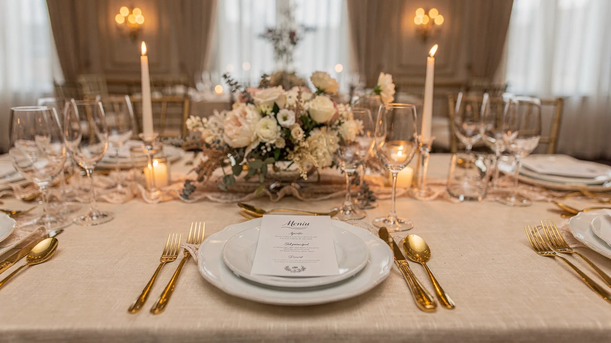 Masă de nuntă elegantă, cu aranjament floral neutru, farfurii albe, tacâmuri aurii, pahare de vin și apă, meniu tipărit și lumânări; atmosfera este rafinată și potrivită pentru un ballroom sau o sală premium.