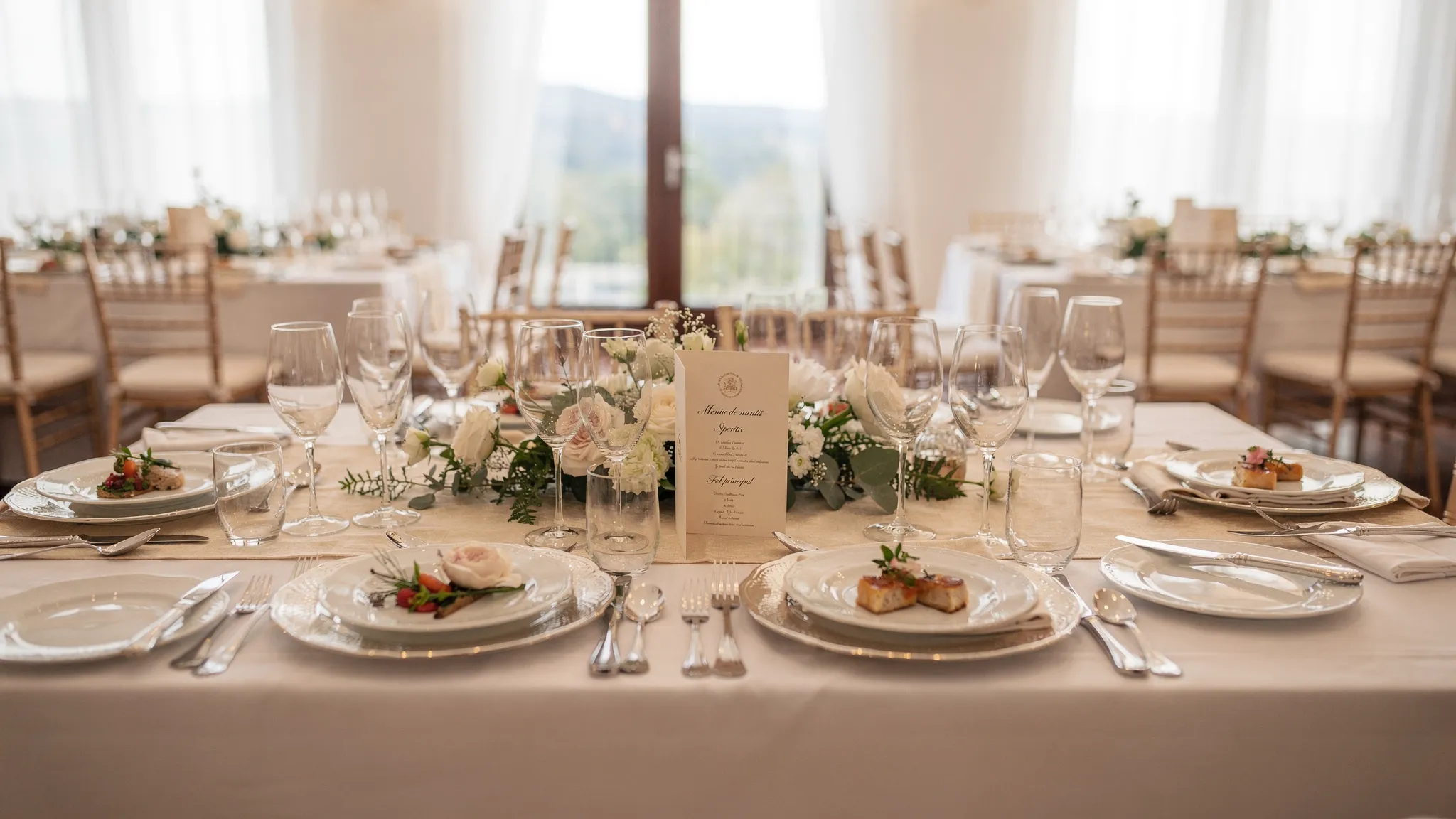 Meniu nuntă Jubile: cum îl personalizezi după invitați 2 O masă elegant aranjată pentru degustarea meniului de nuntă, cu farfurii de prezentare pentru aperitiv și fel principal, carduri de meniu, pahare pentru vin și apă, și elemente florale discrete într-o sală luminoasă de evenimente.