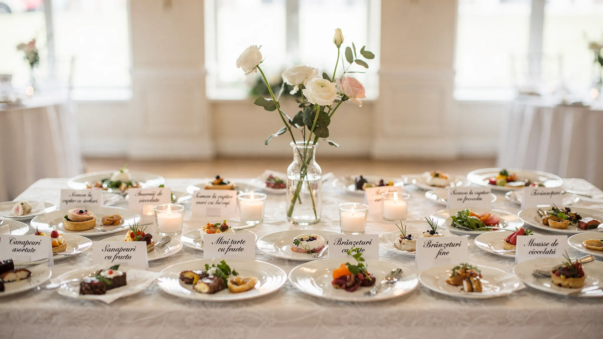O masă elegantă de degustare pentru nuntă, cu farfurii de prezentare în porții mici, etichete discrete pentru fiecare preparat și un aranjament floral minimalist.