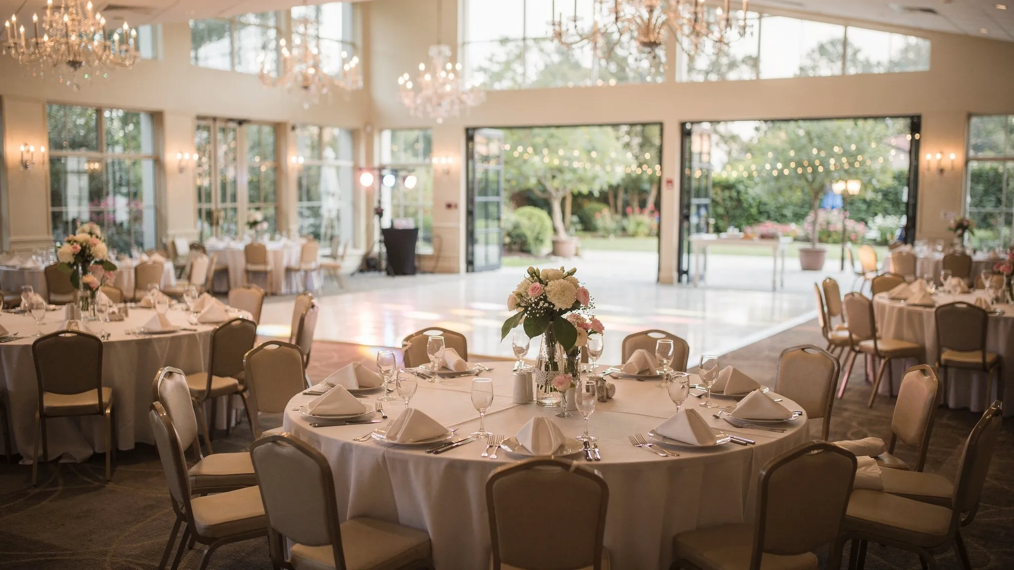 O sală elegantă pentru botez, cu mese rotunde aranjate armonios, aranjamente florale discrete, lumini calde, ring de dans vizibil și acces către o zonă exterioară tip grădină/terasă.