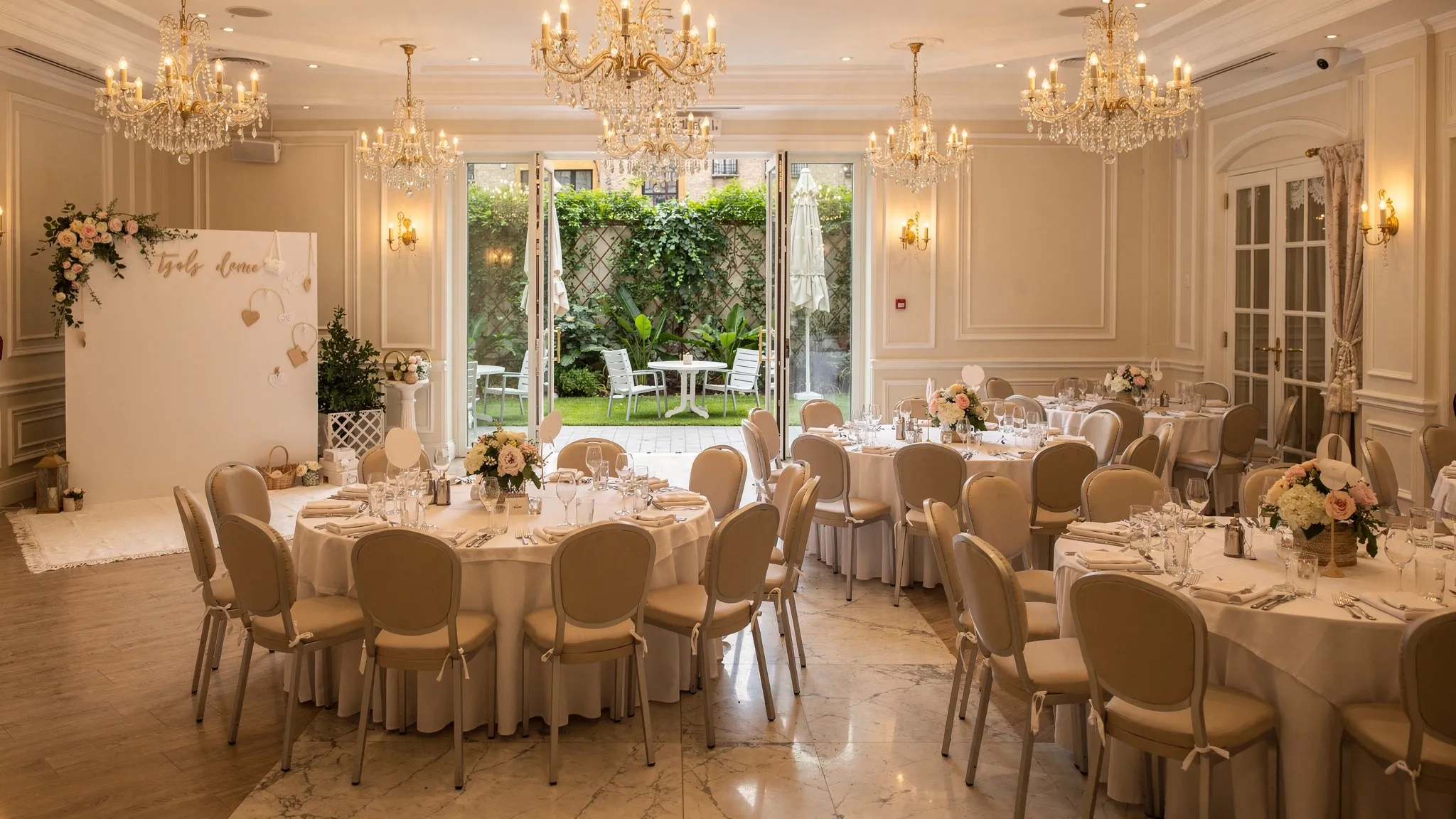 O sală elegantă pregătită pentru botez, cu mese rotunde aranjate impecabil, lumini calde, un colț foto discret și acces vizibil către o terasă verde pentru aer liber.
