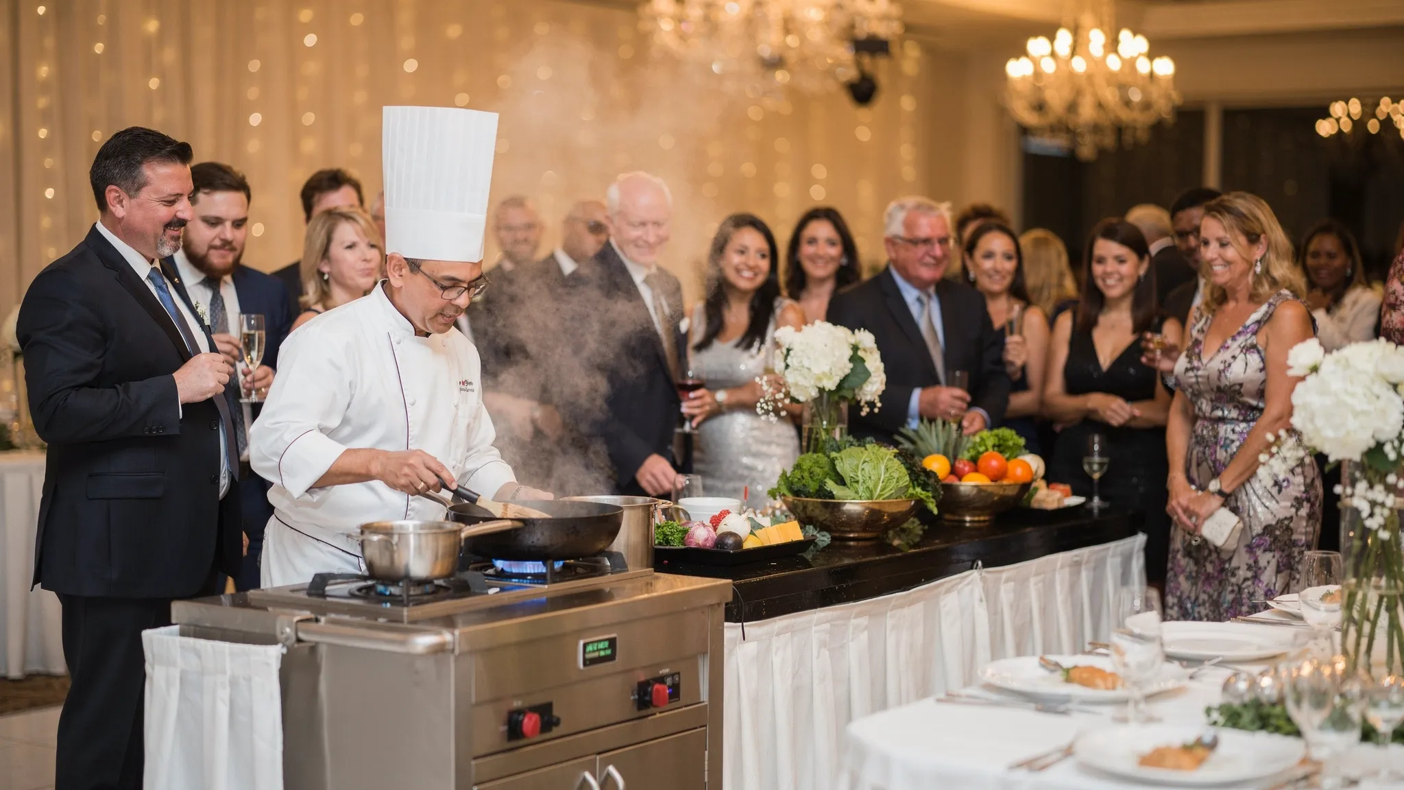 O stație de live cooking la un eveniment, cu bucătar care gătește la vedere și invitați adunați în jur, în decor elegant de nuntă.