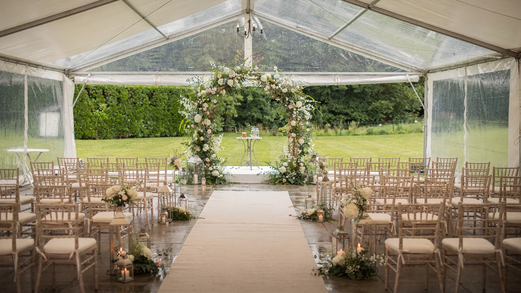 Spațiu acoperit pentru plan B la o cununie în aer liber, cu arc floral montat în fața unei zone luminoase, scaune așezate simetric și covor discret pentru alee, pregătit pentru ploaie.