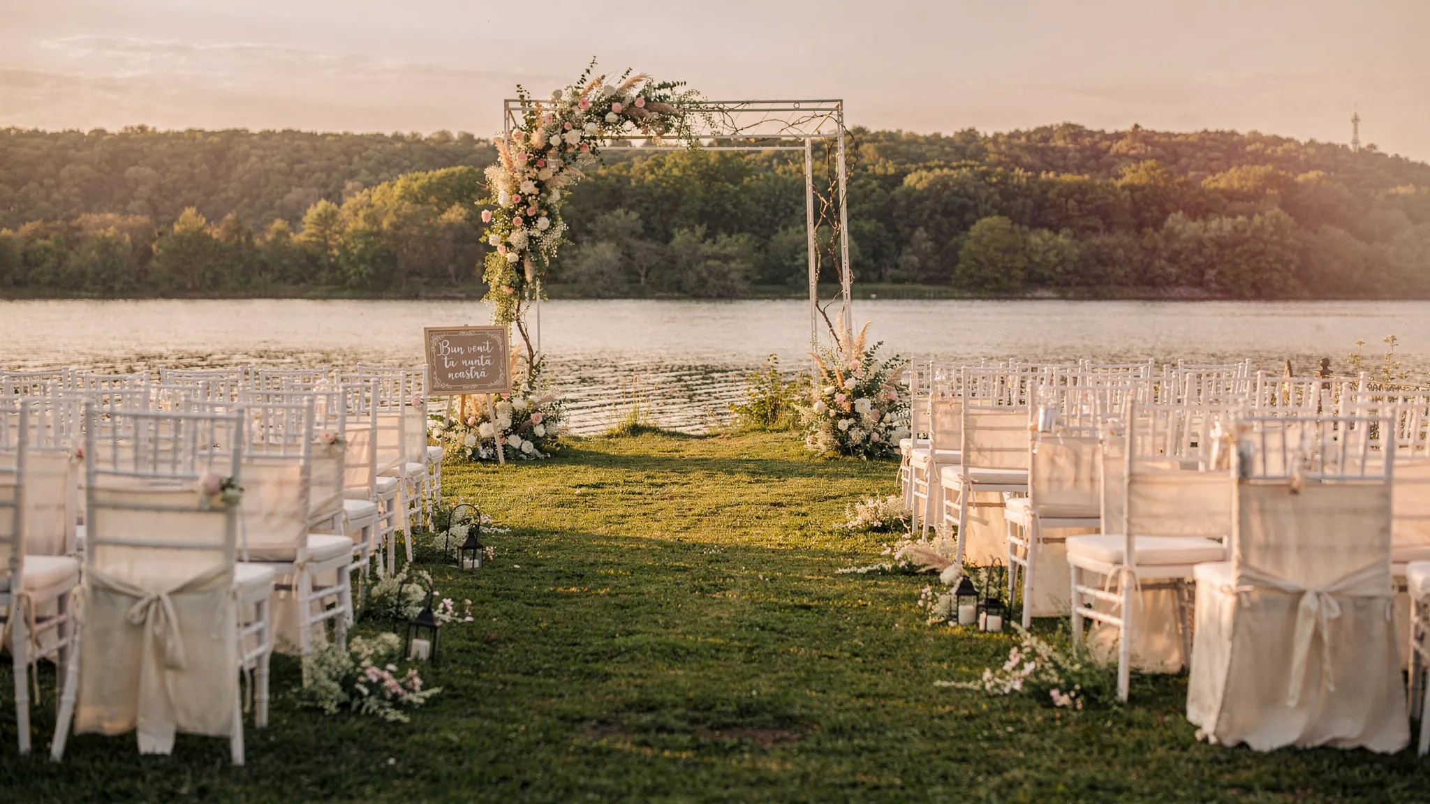Nuntă în București: cum obții vibe de destinație fără drum 2 Ceremonie de nuntă în aer liber lângă București, cu scaune elegante aranjate pe un culoar, pe fundal apă sau pădure, lumină caldă de apus și un arc floral minimalist.