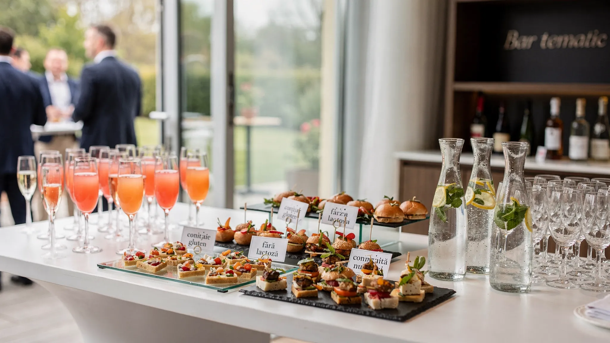 Meniu evenimente: cum calculezi corect cantitățile și momentele 3 O masă de cocktail elegantă la un eveniment, cu platouri de finger food, pahare cu welcome drink, carafe cu apă, un colț de bar tematic și etichete discrete pentru alergeni, într-un spațiu luminos indoor-outdoor.